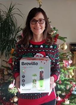 Winner of food blender holds her prize and stands in front of Christmas tree.